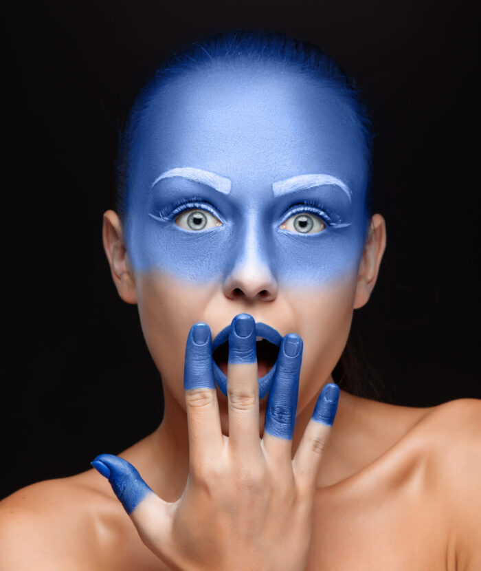 Portrait of a young afraid  woman who is posing covered with blue paint in the studio on a black background. Girl has blue lips and white eyebrows